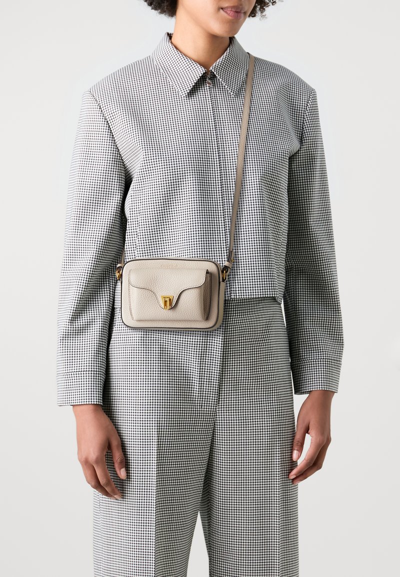 Crossbody-veske i teksturert beige skinn med gullfarget spenne, rektangulær form og kontrastfarget mørk kant, båret over en rutete skjorte.