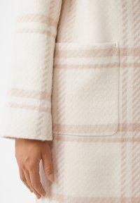Cappotto di lana crema con una consistenza morbida, caratterizzato da un delicato motivo a quadri rosa chiaro e una tasca applicata. La manica ha un taglio leggermente svasato.