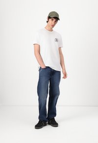 White short-sleeve t-shirt with small graphic, blue denim jeans, and black sneakers, topped with an olive green cap. Casual fit.