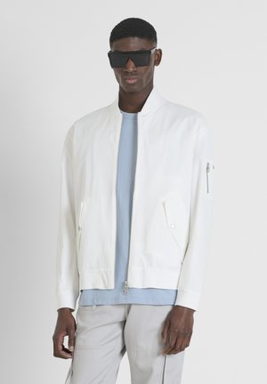 Veste bomber blanche avec une fermeture éclair à l'avant, deux poches avant et une poche zippée sur la manche, portée sur une chemise bleu clair et un pantalon gris.