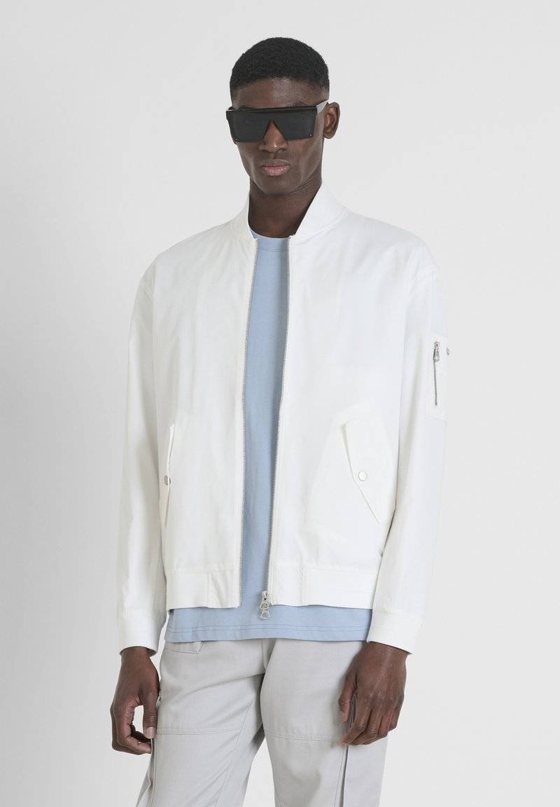 Veste bomber blanche avec une fermeture éclair à l'avant, deux poches avant et une poche zippée sur la manche, portée sur une chemise bleu clair et un pantalon gris.