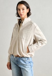 Giacca zip-up beige chiaro con due tasche frontali, realizzata in tessuto testurizzato; indossata sopra una maglietta color sabbia, abbinata a jeans azzurri chiari.