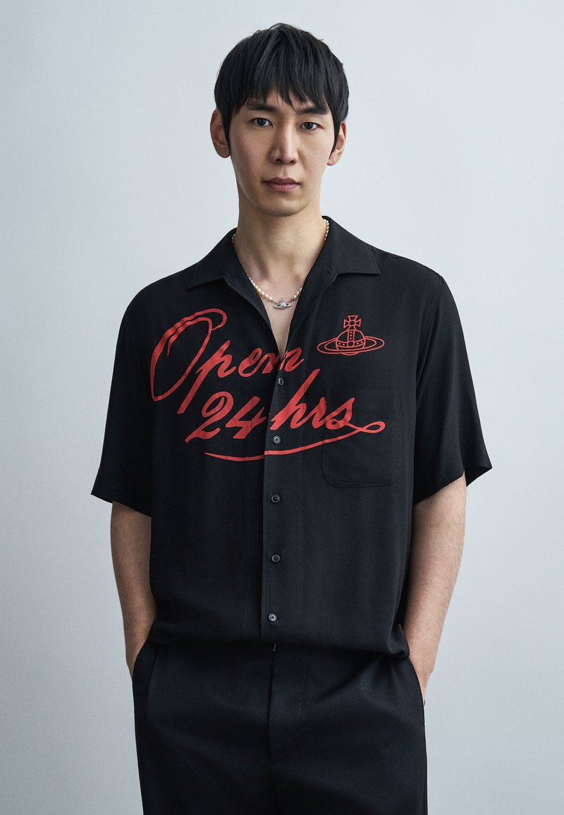 Chemise noire à manches courtes avec un graphisme rouge "Ouvert 24 h" sur le devant. Dotée d'une fermeture à boutons et d'une poche sur le côté gauche. Texture lisse.