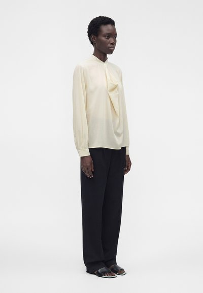 Femme debout portant un chemisier crème à manches longues avec un détail drapé, un pantalon noir à jambes larges et des sandales plates noires ouvertes à l’avant sur fond blanc.