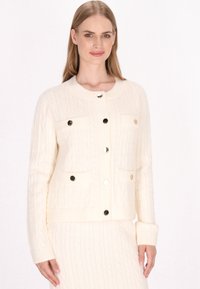 Crème-kleurige kabelgebreide cardigan met twee voorzakken, vier ronde knopen en geribde manchetten; zachte textuur en gestructureerd ontwerp.
