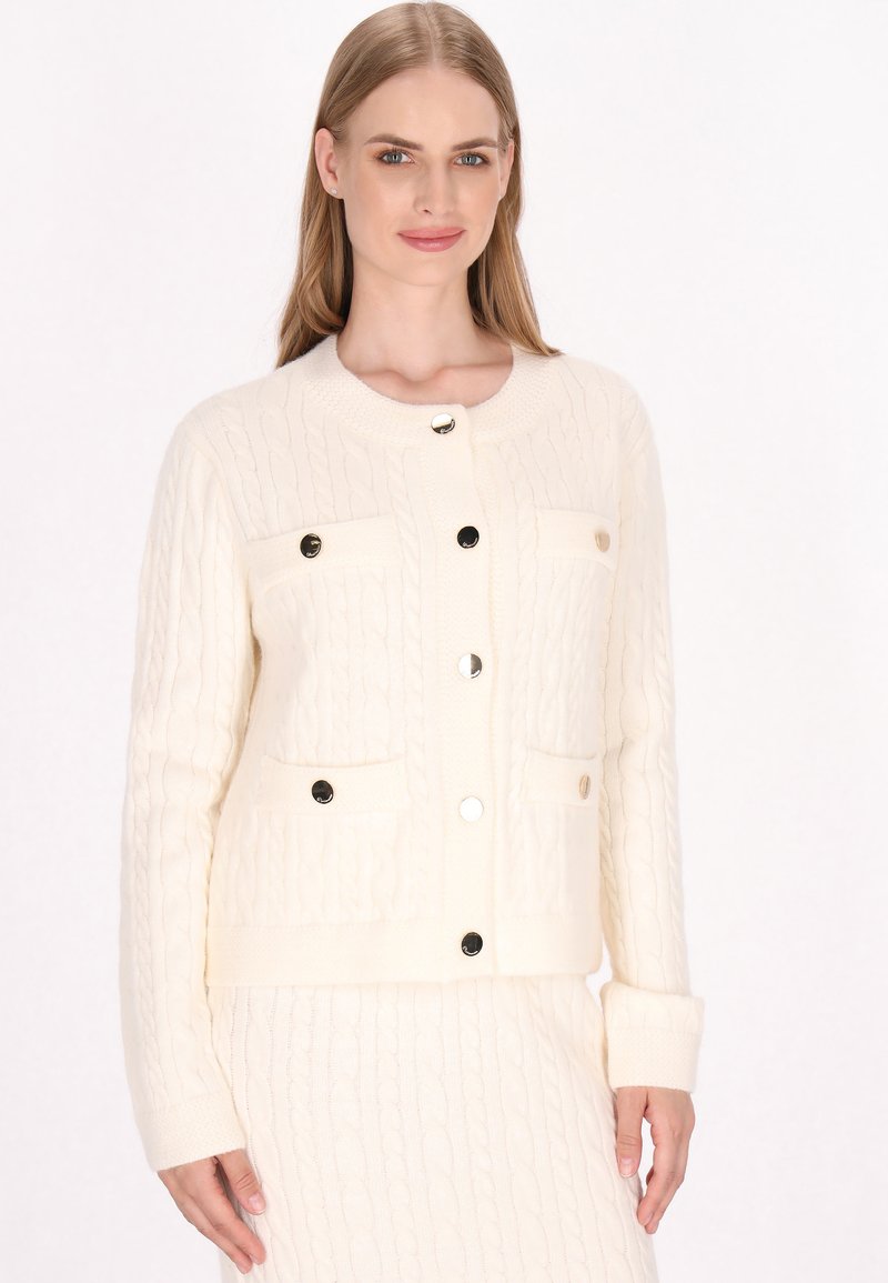 Crème-kleurige kabelgebreide cardigan met twee voorzakken, vier ronde knopen en geribde manchetten; zachte textuur en gestructureerd ontwerp.