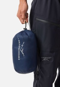 Marinecompressiezak met een trekkoordsluiting, met een wit logo. Vastgehouden in een hand naast donkergekleurde broeken met een ritszak.