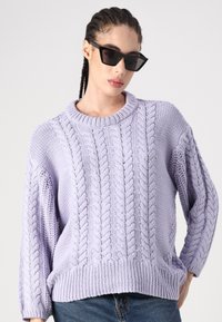 Pull en laine de couleur lavande avec des motifs en relief, un ourlet et des poignets côtelés, et un col ras du cou. Associé à des lunettes de soleil noires angulaires.