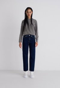 Chemise grise à manches longues, jeans en denim foncé taille haute, et baskets blanches. Design minimaliste, coupe décontractée, sans motifs ni logos visibles.