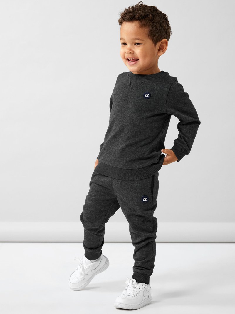 Donkergrijze katoenen sweatshirt en joggers, met ribgebreide boorden en een klein blauw logo op de borst en de broek. Gecombineerd met witte sneakers.