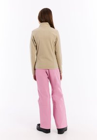Beige fleecejack met een hoge kraag, gecombineerd met roze waterdichte broek en zwarte laarzen, gezien van achteren.