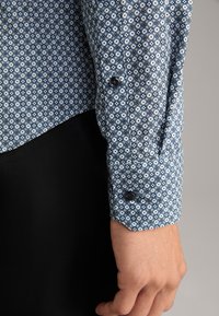 Hellblaues Button-Down-Hemd mit einem geometrischen Muster in dunklerem Blau und Weiß. Verfügt über eine abgerundete Manschette mit schwarzen Knöpfen und einer glatten Textur.