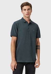 Mørkegrøn polo-shirt lavet af bomuld, med krave, korte ærmer og et lille logo på venstre bryst. Båret med sorte jeans.