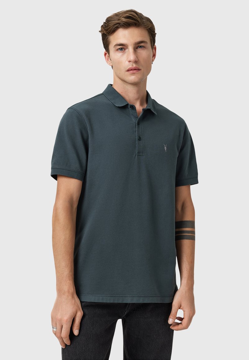 Mørkegrøn polo-shirt lavet af bomuld, med krave, korte ærmer og et lille logo på venstre bryst. Båret med sorte jeans.
