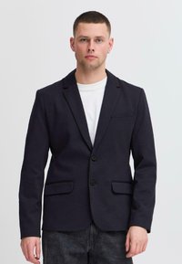 Blazer bleu marine en tissu texturé, dot é d'un col à revers cranté, de deux poches avant et d'une fermeture à un seul bouton. Porté par-dessus une chemise blanche.
