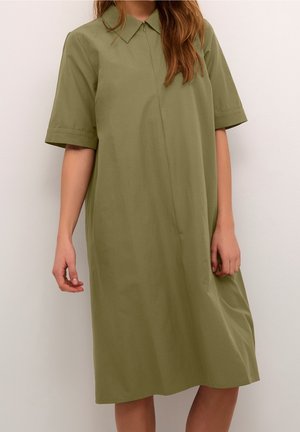 Femme portant une robe-chemise ample, couleur vert olive, arrivant au genou, avec des manches courtes et un col, sur un fond clair uni.