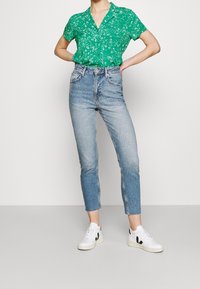 Grünes Blumenhemd mit kurzen Ärmeln, hellblaue, hoch taillierte Jeans in einer verkürzten Länge, kombiniert mit weißen Sneakers mit schwarzen Akzenten.