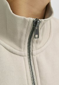Beige zip-up jacka med ribbad krage och textilstruktur. Har en silverfärgad metalldragkedja och synliga sömnadsdetaljer längs sömmarna.