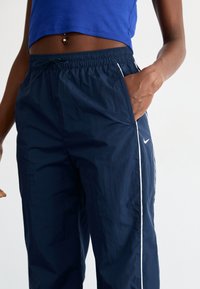 Tengerészkék szélálló nadrág, rugalmas derékkal, fehér oldalcsíkkal és zsebekkel. Könnyű anyagból készült, sima textúrával.