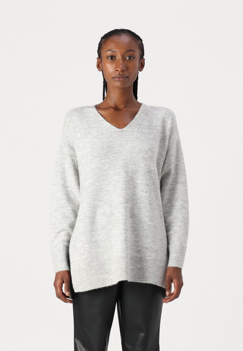 Pull léger gris en V en matière douce. Présente des manches longues et une coupe décontractée, avec un ourlet côtelé et des fentes sur les côtés au bas.