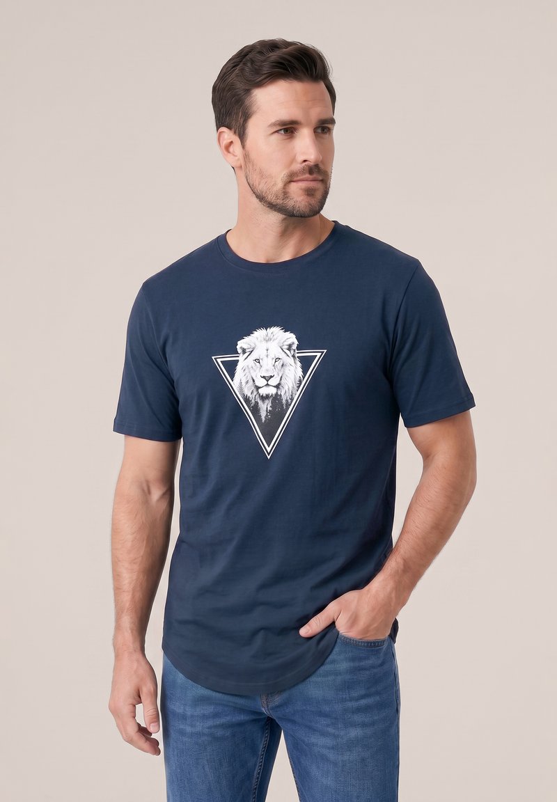 Άνδρας που φοράει μπλε t-shirt με λευκό πρόσωπο λεονταριού μέσα σε τρίγωνο, συνδυασμένο με μπλε τζιν, στέκεται με το ένα χέρι στην τσέπη.