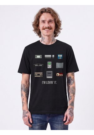Homme avec des tatouages portant un t-shirt noir affichant des consoles et appareils de jeux rétro avec le texte "I'M LOVIN' IT."