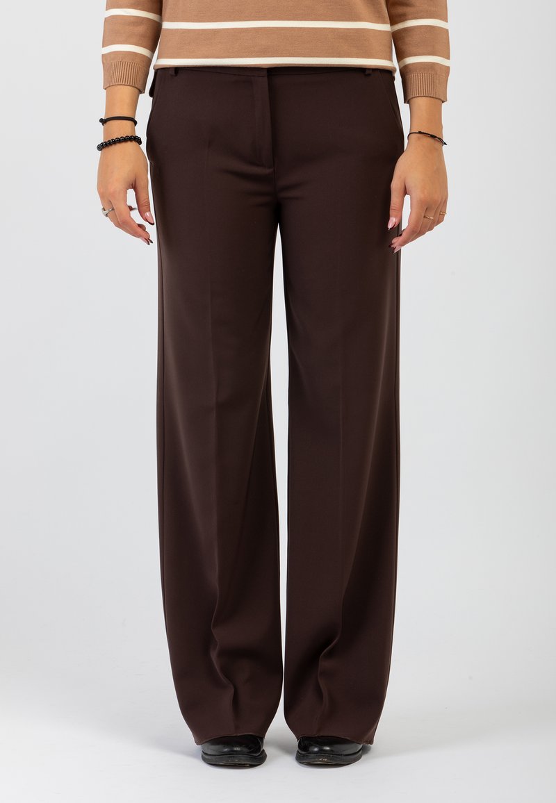 Persona che indossa pantaloni a gamba larga marrone scuro, un maglione a righe marrone chiaro, scarpe nere e braccialetti su entrambi i polsi, in piedi su uno sfondo bianco.