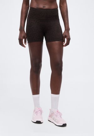 Atletisk person iført sorte tætsiddende shorts, hvide crew-sokker og pink Adidas løbesko står på ensfarvet baggrund.