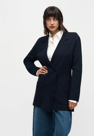 Γυναίκα με σκούρα μαλλιά που φοράει μαύρο φαρδύ σακάκι, λευκό πουκάμισο με στρόγγυλο γιακά και μπλε τζιν με φαρδιά μπατζάκια, στέκεται με το ένα χέρι στη μέση.