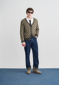 Καφέ και μπεζ καρό ζακέτα, λευκό πουκάμισο με γραβάτα, μπλε τζιν και γκρι μποτάκια. Το μοντέλο φοράει σκουρόχρωμα γυαλιά ηλίου και στέκεται μπροστά σε απλό φόντο.