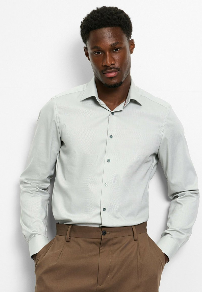 Light gray long-sleeve dress shirt with a button-down collar and subtle texture, paired with brown trousers. Features matching buttons.