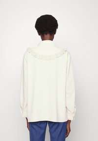 Person with short curly hair wearing a cream long-sleeve blouse with lace trim on the back yoke and blue jeans, facing away.