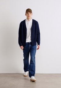 Ναυτικό πλεκτό γιλέκο με κουμπιά, λευκή polo μπλούζα, μπλε τζιν και λευκά αθλητικά παπούτσια. Το μοντέλο περπατάει μπροστά από έναν απλό φόντο.