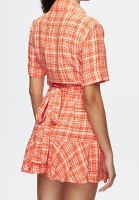 Kortärmad orange rutad klänning med krage, midjeband, utsvängd fåll och mjuk bomullstextur; har vita och mörkare orangea detaljer.