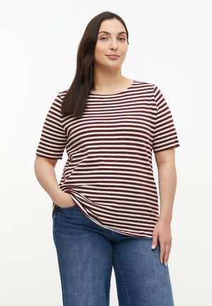 Frau mit langen dunklen Haaren trägt ein kurzärmeliges Shirt mit bordeauxroten und weißen Streifen und blaue Jeans, steht mit einer Hand in der Tasche.