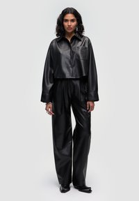 Afgeknot zwart neplederen shirt met knoopsluiting aan de voorkant en borstzak, gecombineerd met losse zwarte neplederen broek en puntige zwarte schoenen.