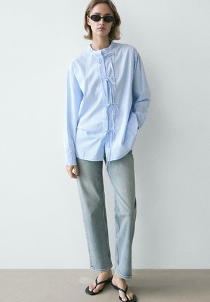 Frau mit schwarzen Sonnenbrillen, lockerem hellblau gestreiftem Hemd mit Knoten vorne, ausgewaschenen Jeans mit geradem Bein und schwarzen Flip-Flops, stehend.