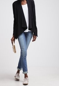 Svart öppen cardigan över en vit skjorta, matchat med ljusblå jeans med uppvikta ärmar och beiga skor, hållande en liten clutch.