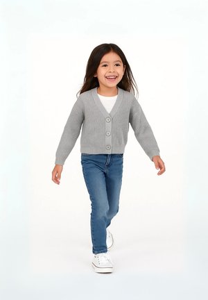 Grå ribbade koftan med fem knappar, bärs över en vit topp, kombinerad med blå jeans och vita sneakers. Barnet går framåt.