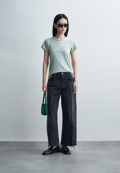 Vrouw met zwarte zonnebril, lichtgroen gestreept T-shirt, wijde donkere jeans, zwarte schoenen, die een kleine groene handtas vasthoudt tegen een effen grijze achtergrond.