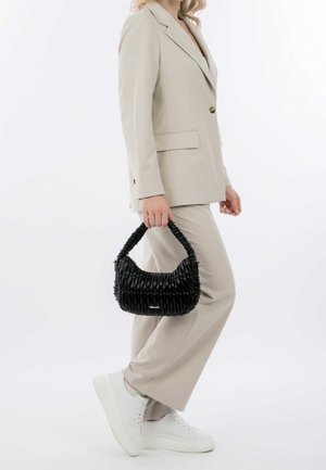 Sort quiltet håndtaske med en buet form og flettet håndtag, båret af en person i en lysebeige dragt og hvide sneakers.