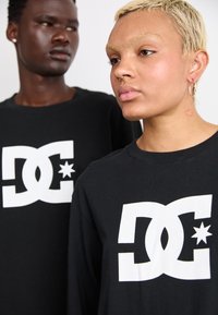 Sorte bomulds t-shirts med et stort hvidt DC-logo med stjernedetaljer, båret af to personer med forskellige frisurer og hudtoner.