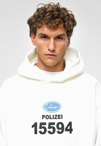 Witte hoodie met een grote zwarte print met "POLIZEI" en het nummer "15594." Zacht materiaal met een comfortabele pasvorm en zichtbare stiksels.