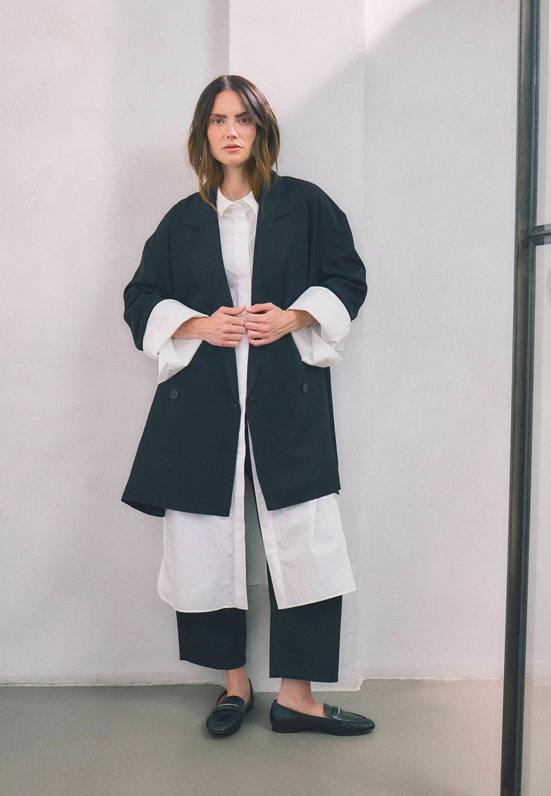 Cappotto oversize nero con polsini bianchi, indossato sopra una camicia bianca con bottoni e pantaloni neri a gamba larga. Abbinato a mocassini neri.