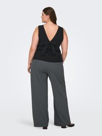 Donna che indossa un top nero senza maniche con scollo profondo a V sul retro e dettaglio a fiocco, abbinato a pantaloni grigi a gamba larga e scarpe scure, in piedi di spalle.