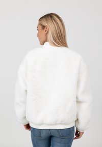 Weiße Kunstfell-Bomberjacke mit hohem Kragen, gerippten Bündchen und Saum; weiche Textur; getragen über blauen Jeans.