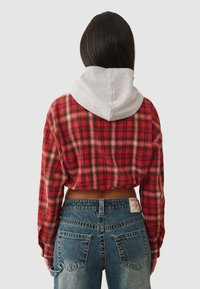 Een persoon met lang zwart haar draagt een korte rode geruite blouse met een grijze capuchon en hooglopende blauwe jeans, met de rug naar de camera gekeerd.