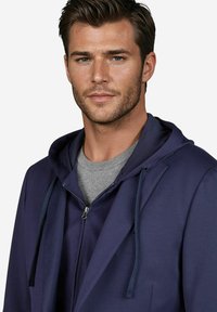 Marineblauwe capuchonjack over een grijze shirt. Heeft een rits aan de voorkant en trekkoorden op de capuchon, en toont een gladde stofstructuur.