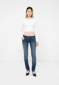 Top blanco recortado de manga larga combinado con unos vaqueros ajustados de tiro medio en azul oscuro. La modelo tiene el cabello lacio y lleva zapatos planos negros de puntera puntiaguda.