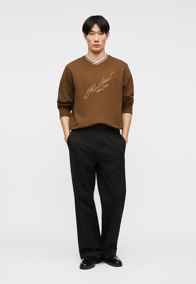 Νέος άνδρας στέκεται με τα χέρια στις τσέπες, φορώντας καφέ μακρυμάνικο πουκάμισο με χειρόγραφο κείμενο, μαύρο παντελόνι και μαύρα παπούτσια σε απλό φόντο.